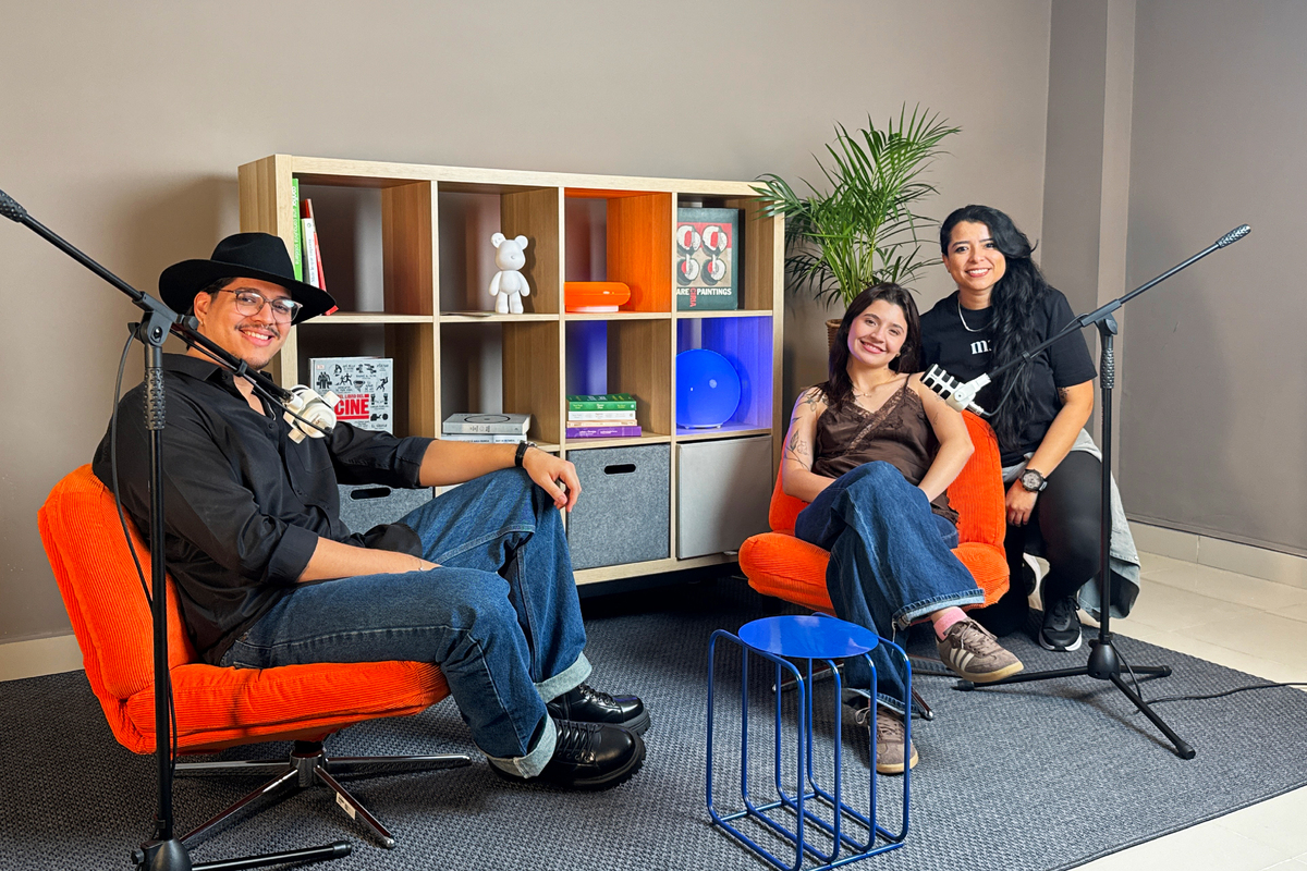 Tres profesionales sonriendo en un estudio de grabación de podcast con micrófonos, sillones naranjas y repisa de fondo.